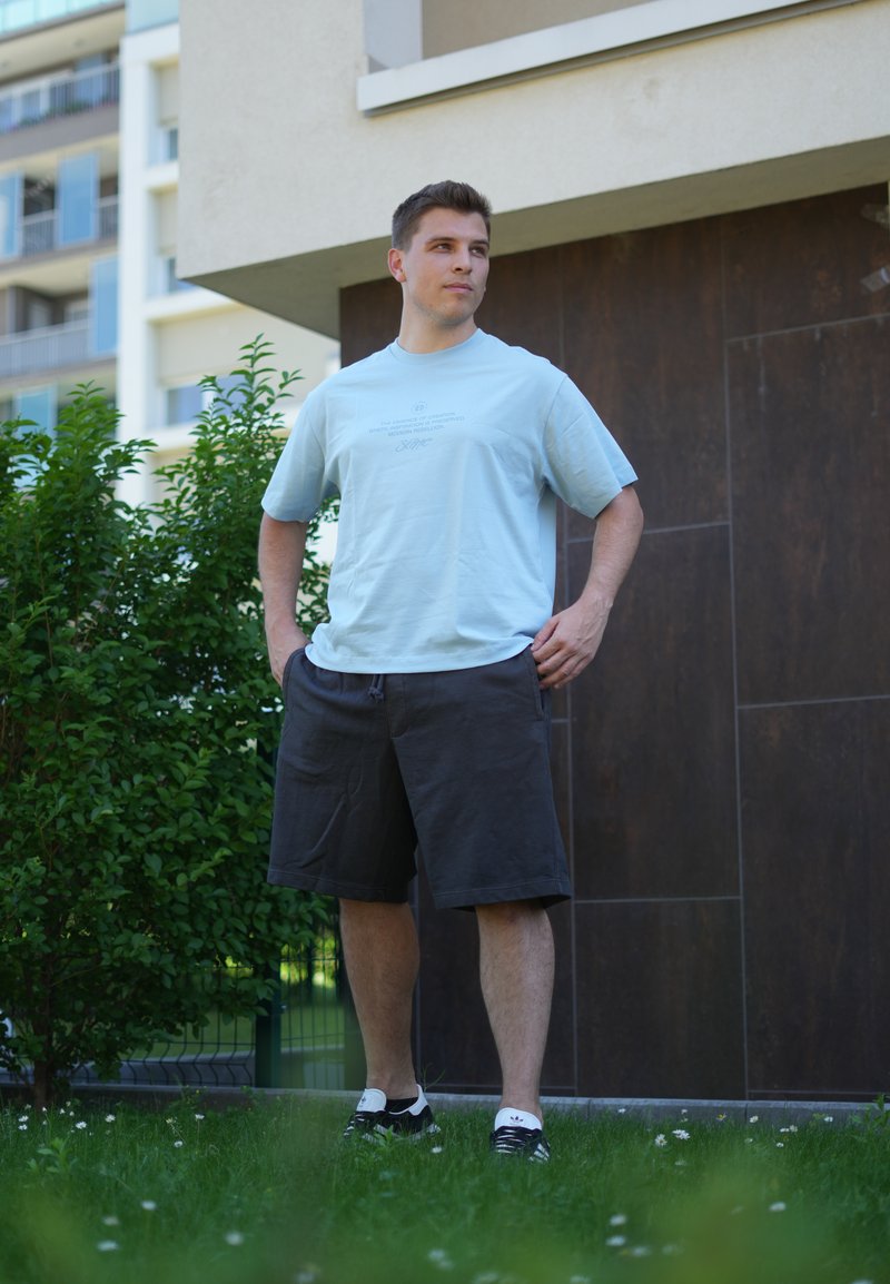 Mann steht draussen auf Gras nahe bei Büschen und einem modernen Gebäude, trägt ein hellblaues T-Shirt, schwarze Shorts und schwarze Sneakers.