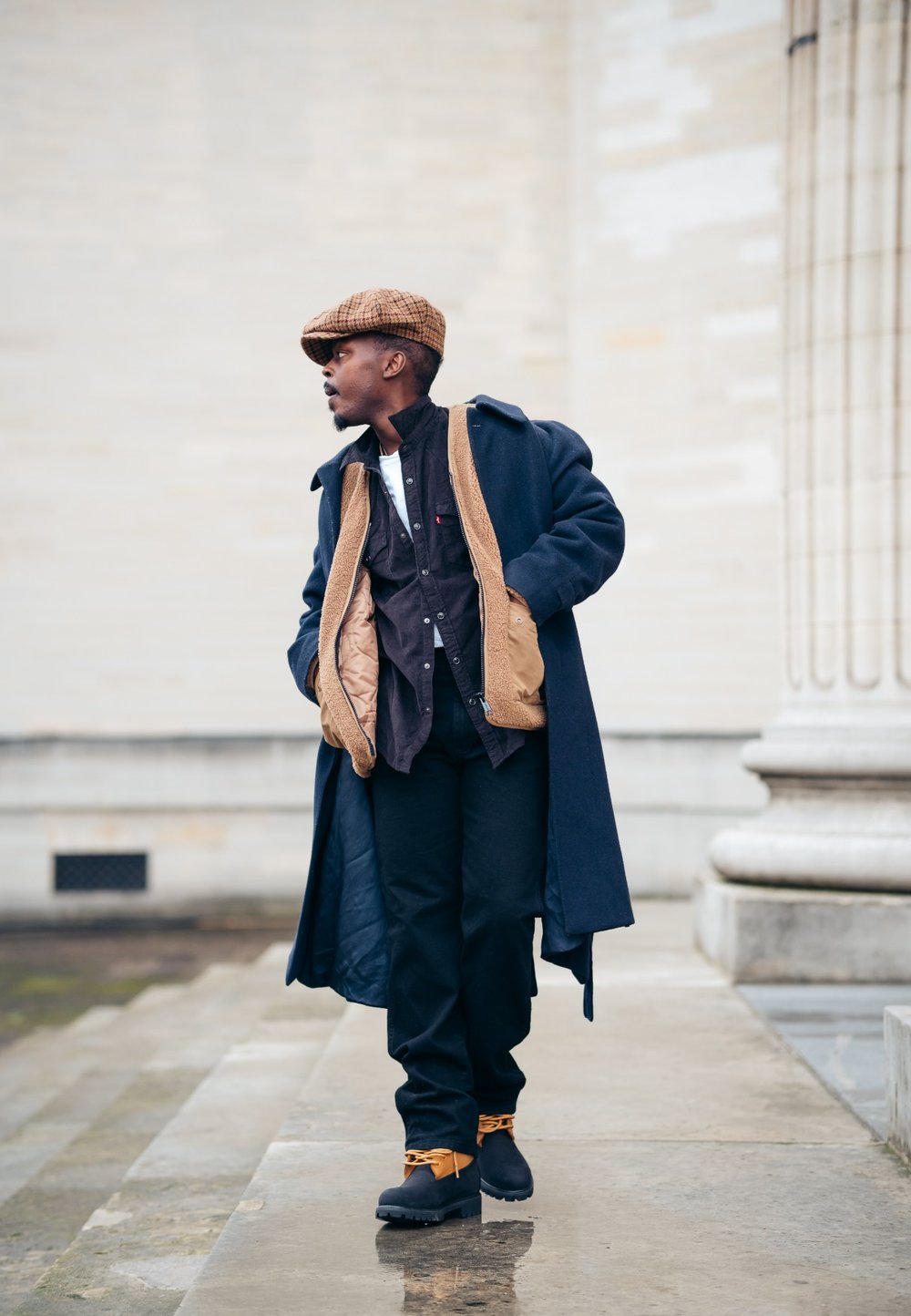 Μακρύ ναυτικό παλτό με καφέ επένδυση, μαύρο πουκάμισο, μαύρο παντελόνι και καφέ καπέλο. Μαύρες ankle μπότες με πορτοκαλί λεπτομέρειες. Αστικός εξωτερικός χώρος.