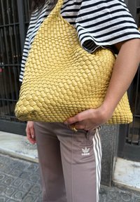 Audzēts dzeltenais rokassomu ar hobo formu, ar gludu tekstūru un lielu, izsistamu dizainu. Lai to turētu, to nes cilvēks, kurš valkā svītrainu augšdaļu un bikses.