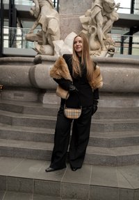 Γυναίκα με μακριά μαλλιά, ντυμένη με μαύρο παντελόνι και σακάκι, με γούνινο μπολερό, μαύρα γάντια και καρό τσάντα, στέκεται σε πέτρινα σκαλιά μπροστά σε μαρμάρινο σιντριβάνι.