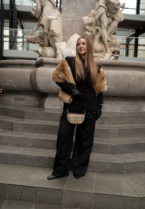 Mujer con cabello largo, vestida con traje de pantalón negro, estola de piel, guantes negros y bolso de cuadros, de pie en escaleras de piedra frente a una fuente de mármol.