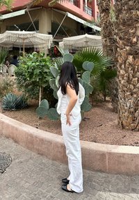 Λευκό, υφασμάτινο ντύσιμο με εφαρμοστό τοπ και φαρδιά παντελόνια. Μαύρες επίπεδες γόβες. Το φόντο περιλαμβάνει φυτά και μ�ια εξωτερική περιοχή καθιστικού.