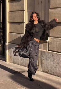 Femme portant un haut noir transparent, une veste duveteuse et un pantalon ample Nike, posant en plein air, en équilibre sur une jambe tout en tenant son autre pied, à côté d'un mur en pierre.