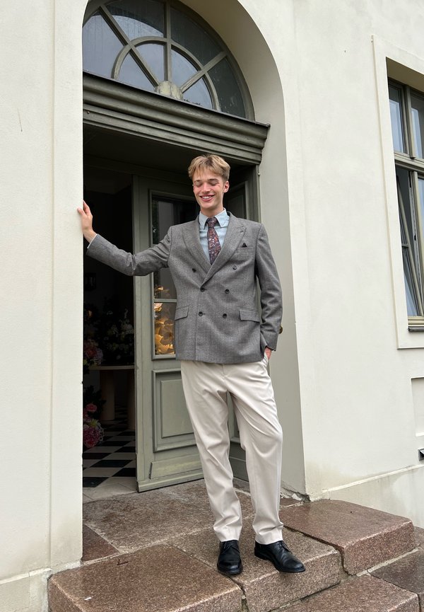Jeune homme souriant, portant un blazer gris, une cravate et un pantalon crème, debout sur des marches en pierre à l'entrée voûtée d'un bâtiment clair.