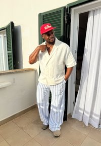 A man wearing a red cap, round glasses, a white short-sleeve shirt, and blue-and-white striped trousers, standing on a balcony beside an open green shutter door.