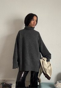 Personne aux cheveux courts et bouclés porte un pull à col roulé gris foncé surdimensionné et un pantalon noir brillant, tenant des vêtements beige et marron.