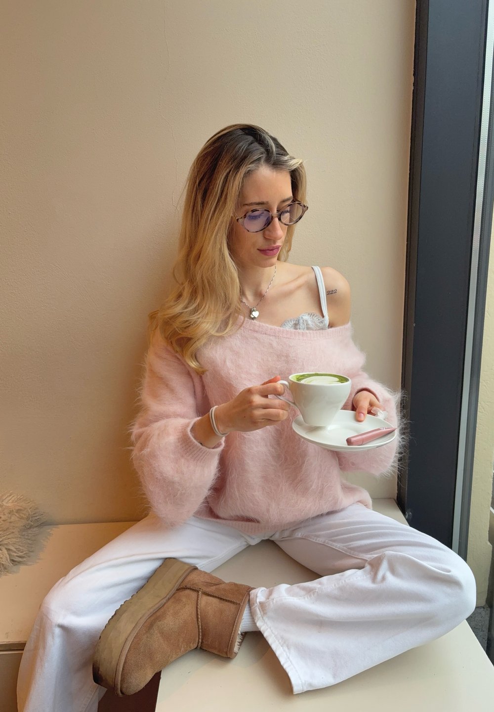 Femme en pull rose pelucheux et pantalon blanc assise en tailleur près de la fenêtre, tenant une tasse blanche avec une soucoupe contenant un latte vert.