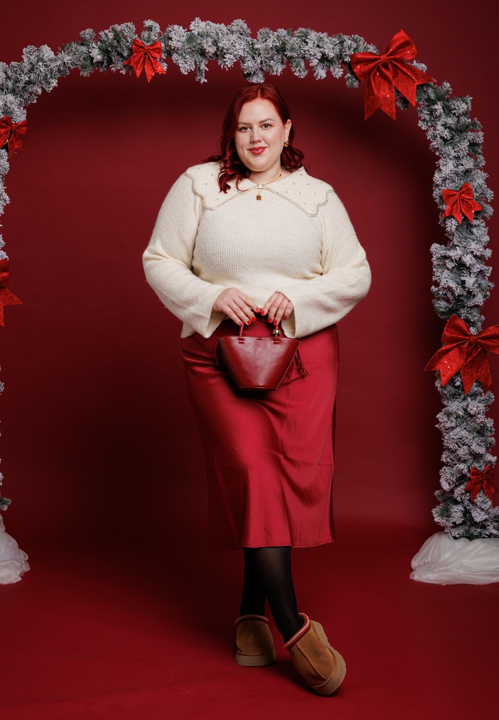 Pull en maille crème avec un col festonné, une jupe en satin rouge, tenant un petit sac à main rouge. Décoré avec une arche festive de verdure et de nœuds.