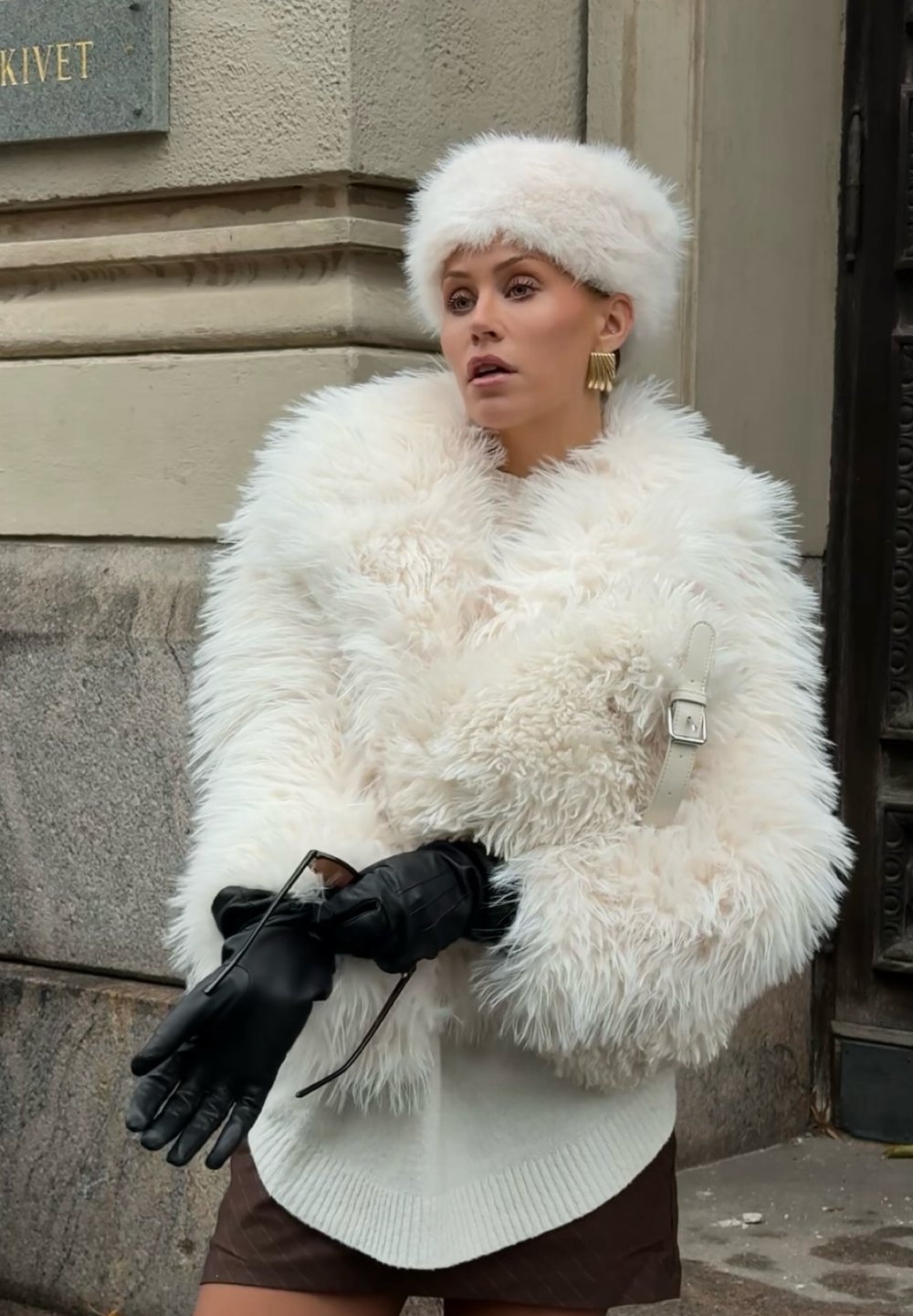 Witte bontjas met een hoge kraag, gecombineerd met een bijpassende bontmuts. Zwarte leren handschoenen en een accessoire riemdetail op de jas.