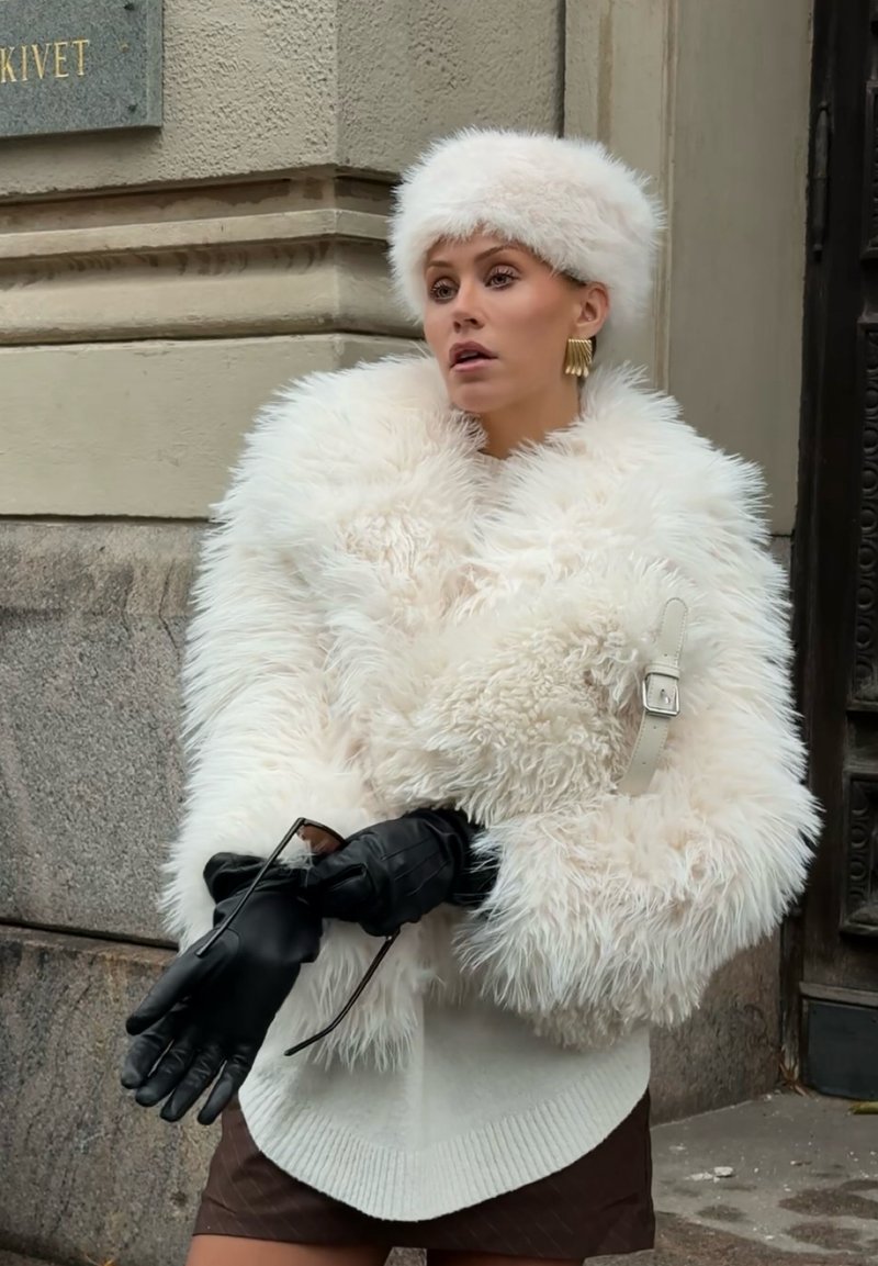 Weißer Pelzmantel mit hohem Kragen, kombiniert mit einem passenden Pelzhut. Schwarze Lederhandschuhe und ein Accessoire-Gürtel-Detail am Mantel.