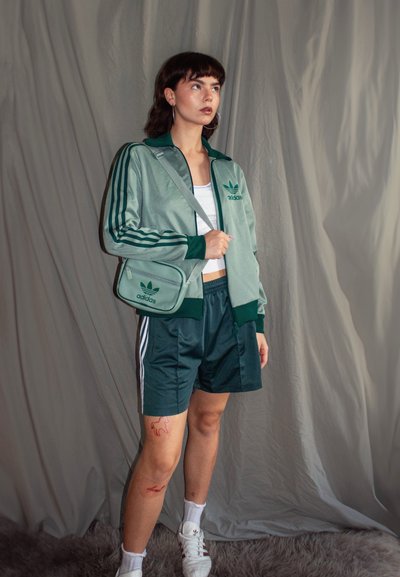 Chaqueta de chándal Adidas verde con acentos negros, pantalones cortos a juego y un bolso de cruzar. Una camiseta sin mangas blanca y zapatillas completan el look deportivo.
