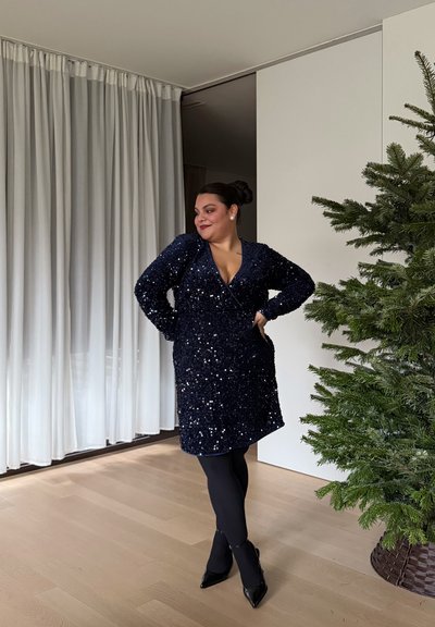 Vestido azul marino con lentejuelas, escote en V y mangas largas, combinado con medias negras y tacones puntiagudos, ambientado frente a un telón y un árbol verde.