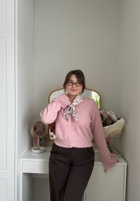 Femme portant des lunettes, un cardigan rose et une écharpe à motifs, debout près d'une coiffeuse blanche avec miroir et bouquet de fleurs.
