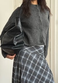 Femme portant un pull gris boutonné et une jupe à carreaux, tenant un grand sac à bandoulière en cuir noir sur un fond uni.