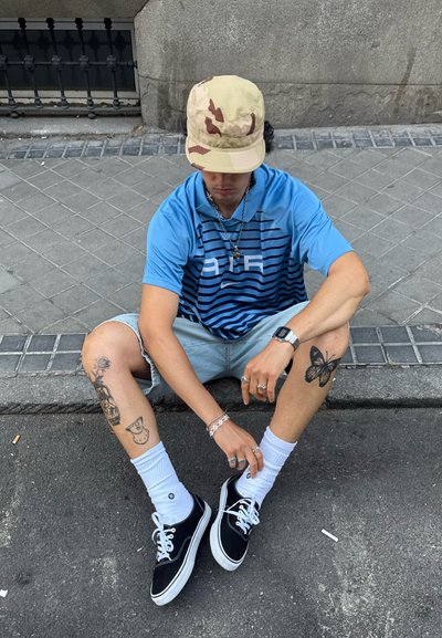 Camisa de manga corta a rayas azules, pantalones cortos de denim claros, zapatillas negras, calcetines blancos y un sombrero tipo bucket de camuflaje claro. Tatuajes visibles en las piernas.