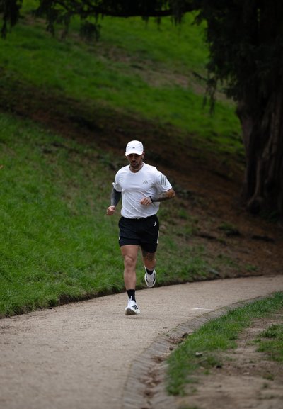 Hombre con gorra blanca, camiseta blanca y pantalones cortos negros corriendo por un camino de grava curvado rodeado de hierba y árboles.