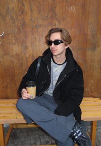 Juodas su užtrauktuku švarkas dėvimas ant pilkos kamufliažo spalvos marškinėlių, pilkos kelnės ir juodi sportbačiai. Laiko rudą kavos puodelį. Medinis fonas ir suoliukas.