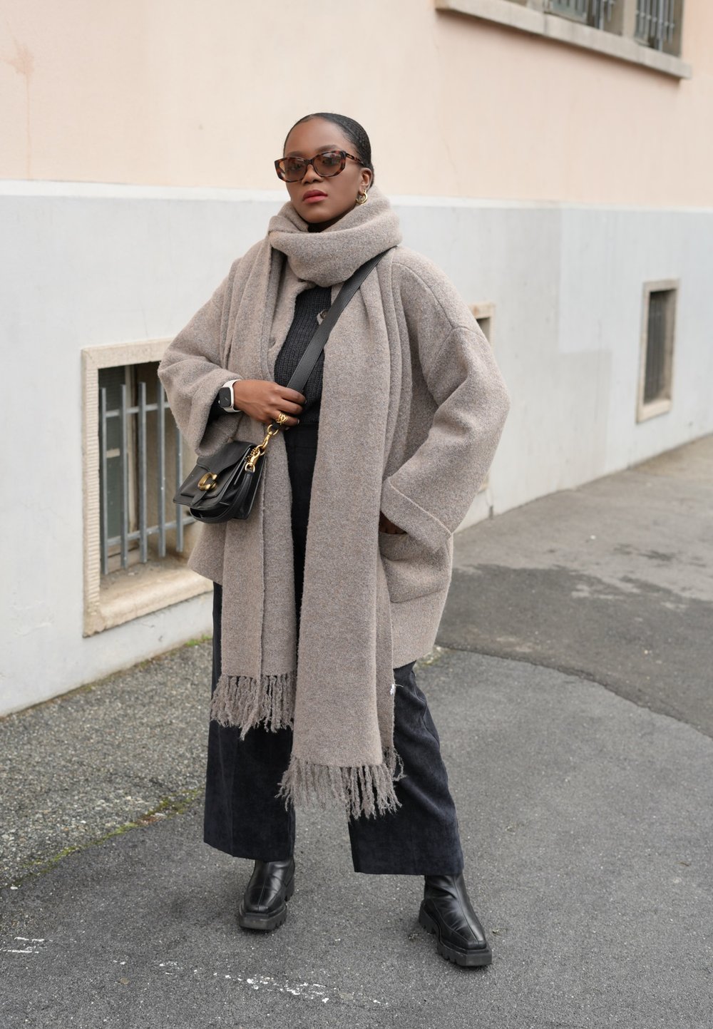 Υπερμεγέθη μπεζ παλτό από μάλλινο υλικό με τσέπες, σε συνδυασμό με ένα ταιριαστό κασκόλ. Μαύρο παντελόνι σε φαρδιά γραμμή και χοντρές μαύρες μπότες μέχρι τον αστράγαλο ολοκληρώνουν το σύνολο.