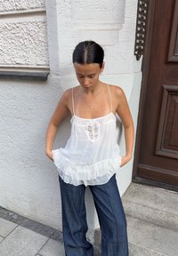 Balta, permatoma liemenė su raukšlėta apatine dalimi ir nėrimo detalėmis, derinama su tamsiomis džinsinėmis pla�čiomis kelnėmis. Modelis laiko rankas kišenėse.