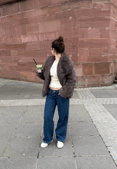 Mujer con chaqueta de piel gris y jeans azules sosteniendo una bebida con pajita, de pie en una calle pavimentada junto a una gran pared de ladrillo rojo.