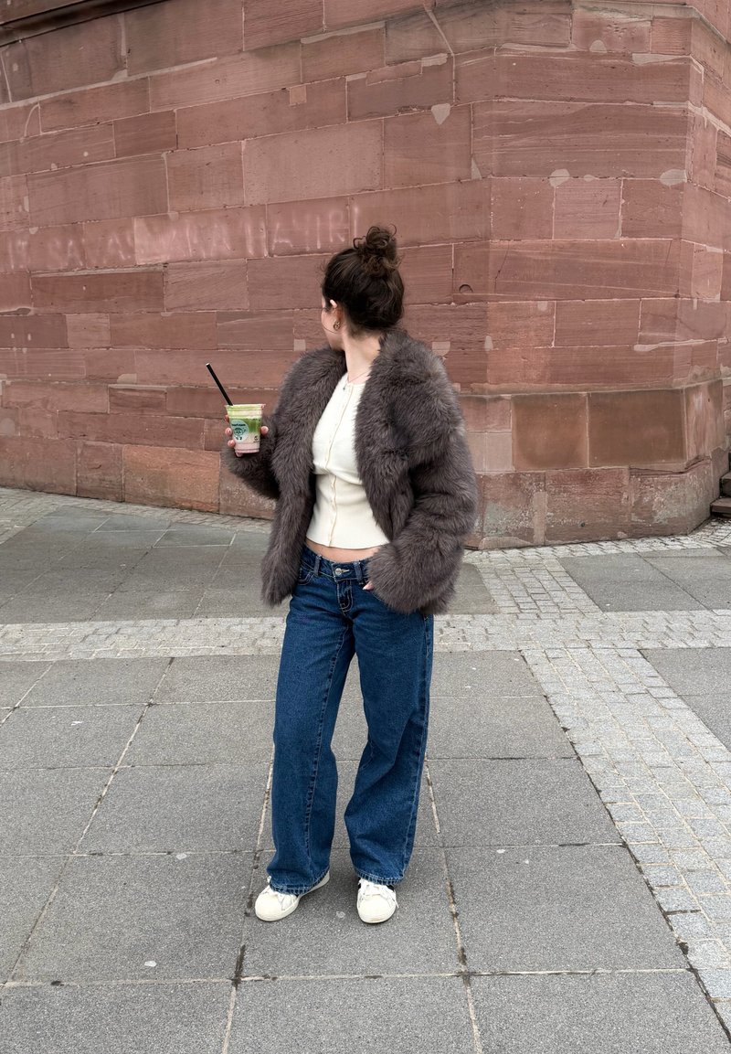 Vrouw in een grijze bontjas en blauwe jeans die een drankje met een rietje vasthoudt, staand op een geplaveide straat naast een grote rode bakstenen muur.