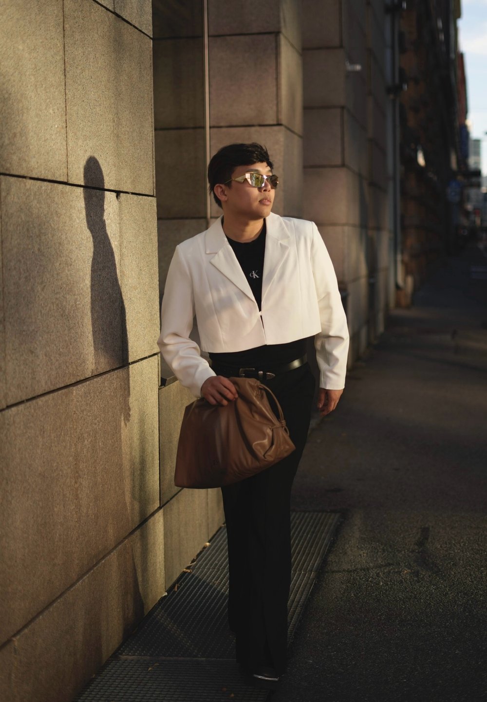 Λευκό κοντό μπουφάν πάνω από μαύρο πουκάμισο, μαύρο παντελόνι, κρατώντας καφέ τσάντα. Λείες υφές, σύγχρονος σχεδιασμός, ανακλαστικά γυαλιά ηλίου.