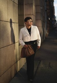 Λευκό κοντό μπουφάν πάνω από μαύρο πουκάμισο, μαύρο παντελόνι, κρατώντας καφέ τσάντα. Λείες υφές, σύγχρονος σχεδιασμός, ανακλαστικά γυαλιά ηλίου.