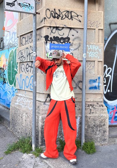 Persona con pantalones y chaqueta naranja ajustándose la gorra, de pie cerca de una pared cubierta de grafitis y un cartel de calle que dice "project district" en una esquina urbana.