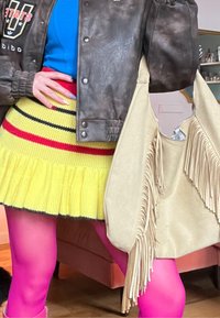 Personne portant une veste marron, un haut bleu, une jupe en maille rayée jaune, des collants rose vif, tenant un sac à franges crème à l'intérieur.