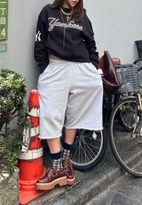 Personne portant un sweat-shirt noir des Yankees, un short large gris, des chaussettes à motifs et des chaussures compensées marron, debout à côté d'un vélo et d'un cône de signalisation orange.