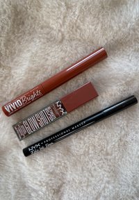 Trzy produkty NYX Professional Makeup: brązowy płynny eyeliner Vivid Brights, bronzowy tint do ust i twarzy Glow Shots, czarny liner Epic Ink na miękkim beżowym tle.