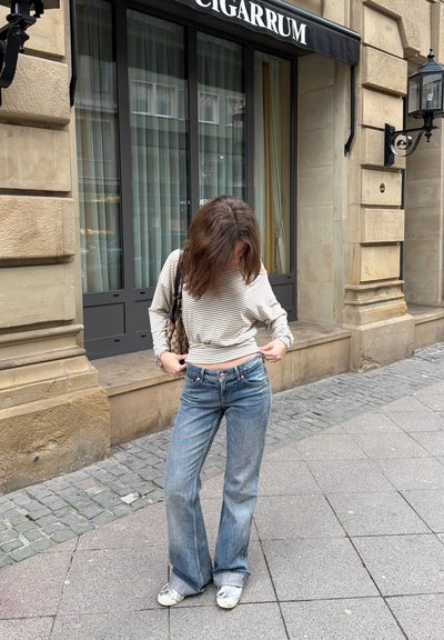 Mujer ajustándose su suéter de rayas mientras está de pie en una acera de la ciudad frente a un edificio de piedra con ventanas grandes y un letrero que dice "CIGARRUM".