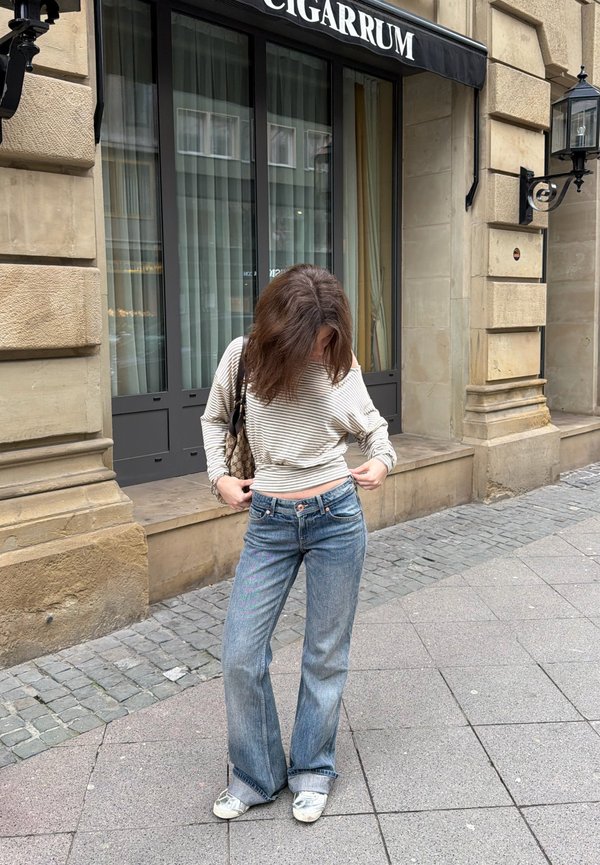 Femme ajustant son pull rayé tout en se tenant sur un trottoir de la ville devant un bâtiment en pierre avec de grandes fenêtres et une enseigne "CIGARRUM".