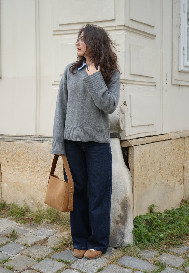 Grigia maglione a maglia con polsini a coste, abbinato a jeans a gamba larga blu scuro. Borsa a spalla marrone e sneakers beige. Sfondo in pietra strutturata.