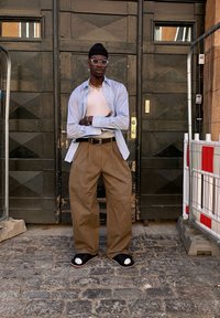 Šviesiai mėlyna marškiniai su mygtukais, rudi plačių kojų kelnės, juodi šlepetės su baltomis akcentais, auksiniai papuošalai, stovintys prieš tamsiai medin door.