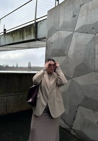 Beige blazer, ribbet lys grå nederdel, transparente solbriller og en struktureret brun håndtaske mod en geometrisk betonvæg og overskyet himmel.