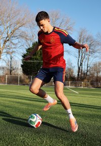 Jeune homme en tenue de football espagnol dribblant un ballon sur un terrain herbeux avec des arbres sans feuilles et un ciel dégagé en arrière-plan.