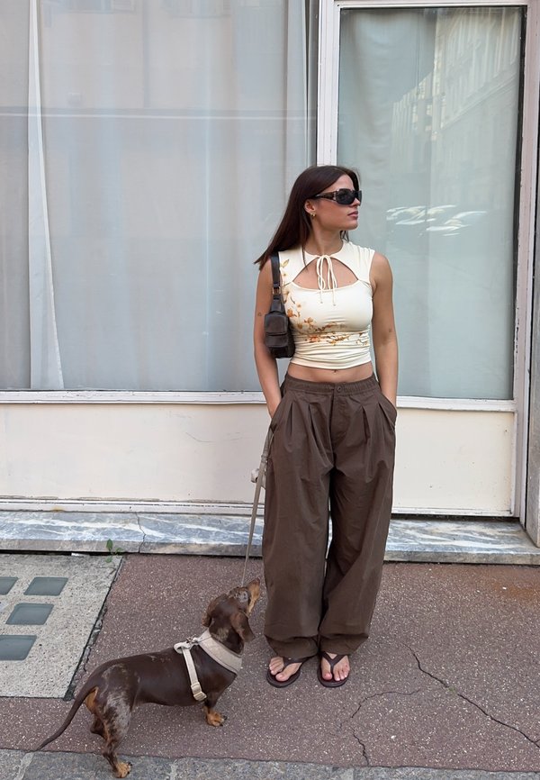 Vrouw met zonnebril die een crèmekleurige crop top en losse bruine broek draagt, staat op het trottoir en houdt een kleine bruine hond aan de lijn.