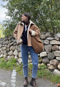 Kvinne i beige lammeskinnsjakke, marineblå genser, blå jeans og brune støvler står ved en steinmur, og bærer en stor brun totebag.