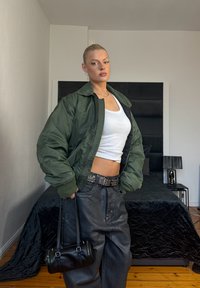 Kobieta w zielonej bomberce, białym krótkim topie, czarnych skórzanych spodniach z paskiem z ćwiekami, trzymająca czarną torebkę, stoi w sypialni.