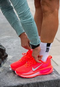 Biegańskie buty Nike ZoomX w jaskrawym pomarańczowym kolorze z srebrnymi akcentami, z teksturowaną cholewką i amortyzującą podeszwą, są właśnie sznurowane.