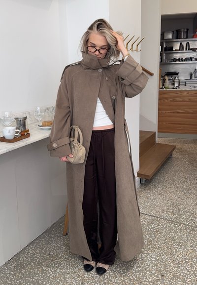 Joven mujer con gafas se ajusta el cabello, lleva un abrigo largo marrón, top corto blanco, pantalones oscuros, sosteniendo un pequeño bolso beige en una cocina moderna.