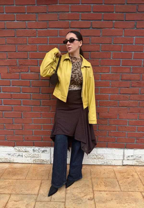 Woman wearing yellow jacket, leopard print top, asymmetrical brown skirt over jeans, black pointed shoes, and sunglasses against red brick wall.