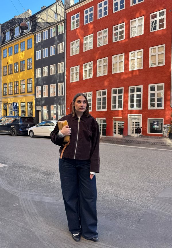 Young woman holding a tan bag stands on a city street with colorful yellow, black, and red multi-story buildings in the background.