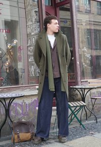 Casaco longo verde de lã, colete com padrão, t-shirt branca, calças largas de ganga azul, botas de couro castanhas, de pé junto a mesas num ambiente urbano.