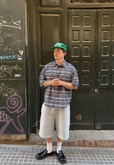 Camisa de cuadros en gris y negro, pantalones cortos grises, zapatos negros, calcetines blancos y gorra verde. El fondo presenta puertas verdes y graffiti.