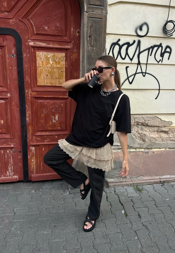 T-shirt oversized preto, saia em camadas bege com renda, calças risca de giz, sandálias pretas, segurando um copo, colar de corrente prateada, óculos de sol.