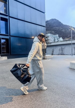 Hombre con chándal gris y gorra caminando afuera cerca de un edificio moderno, llevando una gran bolsa de viaje negra al anochecer.