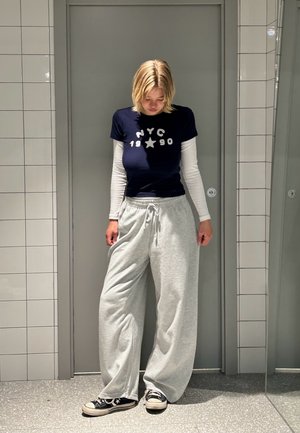 Marinefarbenes T-Shirt mit weißem NYC 1990 Aufdruck, übereinander getragen mit einem langen weißen Oberteil. Graue Jogginghosen und schwarze Sneakers vervollständigen den lässigen Look.