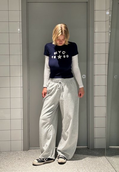 Camiseta navy con impresión blanca de NYC 1990, superpuesta sobre una camiseta blanca de manga larga. Pantalones de chándal grises y zapatillas negras completan el look casual.